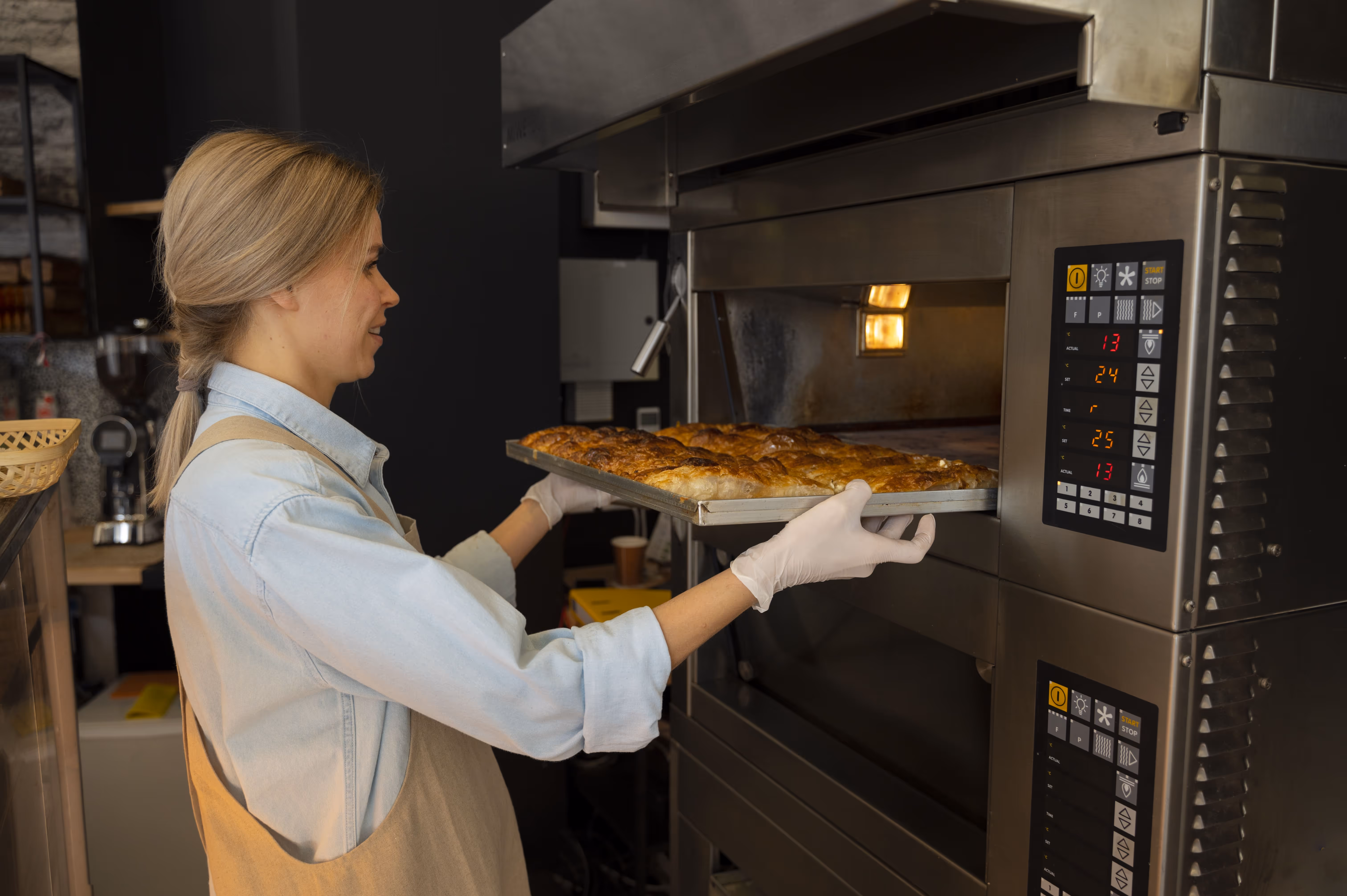 Mulher tirando uma assadeira com pães dourados de um forno industrial em padaria.