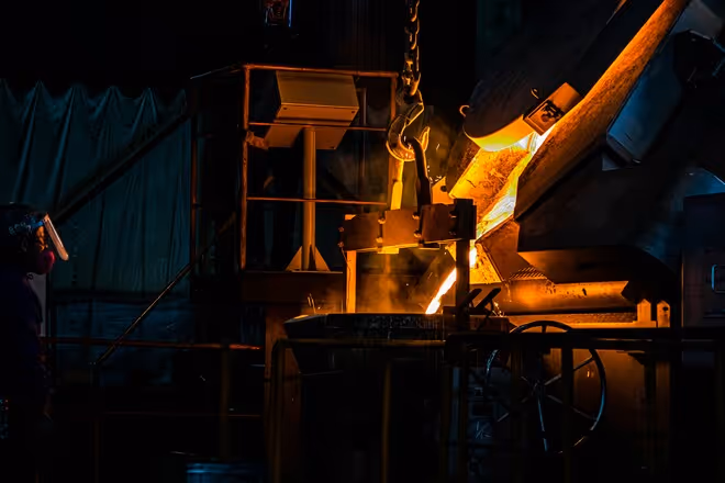 Trabalhador usando máscara enquanto acompanha o derramamento de metal fundido em uma metalúrgica com iluminação escura e quente.