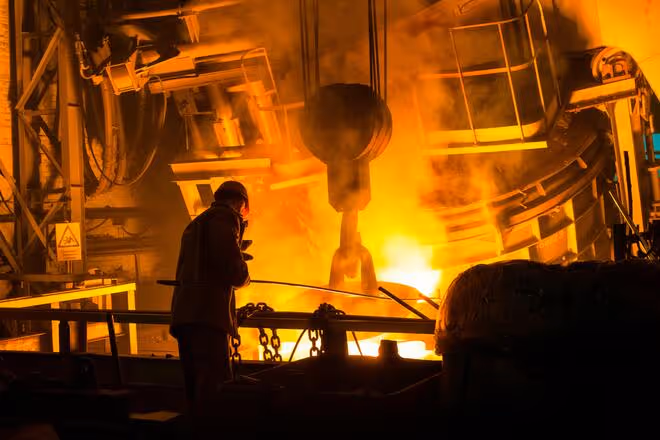 Trabalhador em metalúrgica operando próximo a forno de fundição com chamas intensas e equipamento de elevação.